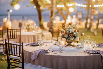 Wedding banquet or gala dinner decorated with garlands. Round tables styled with flowers, candles and accessories. Open air festive banquet on green lawn during dusk