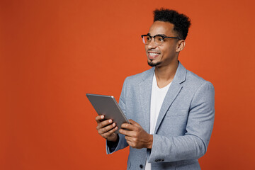 Wall Mural - Young employee business man corporate lawyer wears classic formal grey suit shirt glasses work in office use digital tablet pc computer look aside area isolated on plain red orange background studio.