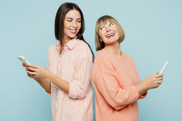 Wall Mural - Side view elder parent mom with young adult daughter two women together wear casual clothes hold in hand use mobile cell phone stand back to back isolated on plain blue background. Family day concept.