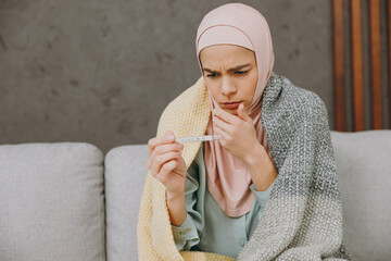 Wall Mural - Young sick muslim woman wear hijab casual clothes plaid check thermometer sits on sofa stay at home flat rest relax spend free spare time in living room indoor People middle eastern uae islam concept.