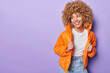 Indoor shot of positive thoughtful woman with curly hair smiles broadly looks aside dressed in outerwear feels pleased isolated over purple background copy space for your promotional content
