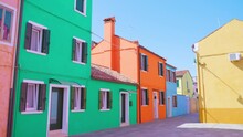 Quiet Manless Streets With Multi-colored Semi-detached Houses On Shady Side And Lit By Morning Sunlight Under Blue Sky In Burano
