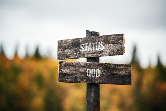 Wall Mural - vintage and rustic wooden signpost with the weathered text quote status quo, outdoors in nature. blurred out forest fall colors in the background.