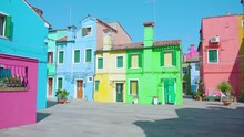 Rows Of Multi-colored Semi-detached Houses Stand On Manless Street Under Blue Cloudless Sky And Bright Sun Rays In Early Morning In Burano