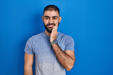 Sticker - Middle east man with beard standing over blue background touching mouth with hand with painful expression because of toothache or dental illness on teeth. dentist