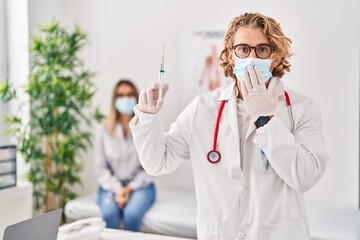 Poster - Blond man wearing doctor uniform and medical mask holding syringe covering mouth with hand, shocked and afraid for mistake. surprised expression