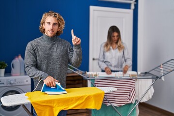 Poster - Couple ironing clothes at laundry room surprised with an idea or question pointing finger with happy face, number one