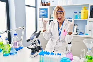 Canvas Print - Middle age blonde woman working at scientist laboratory shouting with crazy expression doing rock symbol with hands up. music star. heavy music concept.