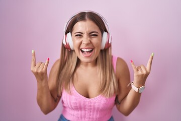 Poster - Young blonde woman listening to music using headphones shouting with crazy expression doing rock symbol with hands up. music star. heavy music concept.