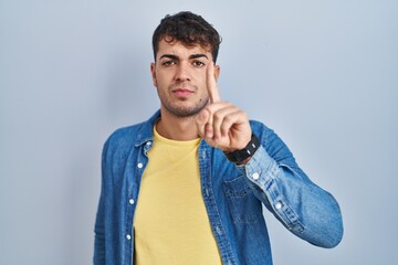 Sticker - Young hispanic man standing over blue background pointing with finger up and angry expression, showing no gesture