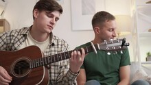 Music Band. Male Composer. Home Leisure. Inspired Happy Two Men Playing Guitar And Singing Sitting Sofa In Light Room Interior.