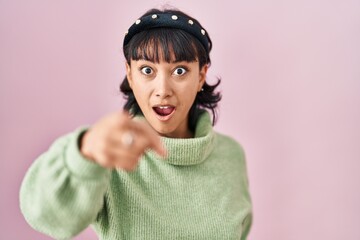 Young beautiful woman standing over pink background pointing with finger surprised ahead, open mouth amazed expression, something on the front