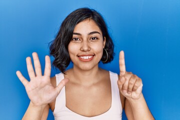 Poster - Young hispanic woman standing over blue background showing and pointing up with fingers number six while smiling confident and happy.