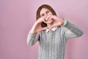 Poster - Beautiful woman standing over pink background smiling in love doing heart symbol shape with hands. romantic concept.