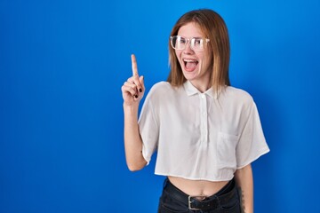 Sticker - Beautiful woman standing over blue background pointing finger up with successful idea. exited and happy. number one.