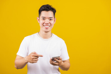 Wall Mural - Asian handsome young man smiling positive holding smartphone blank screen and pointing finger to mobile phone, studio shot isolated on yellow background, making successful expression gesture concept