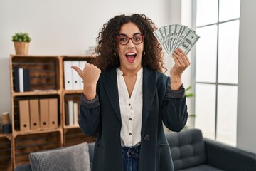 Sticker - Young hispanic woman holding dollars pointing thumb up to the side smiling happy with open mouth