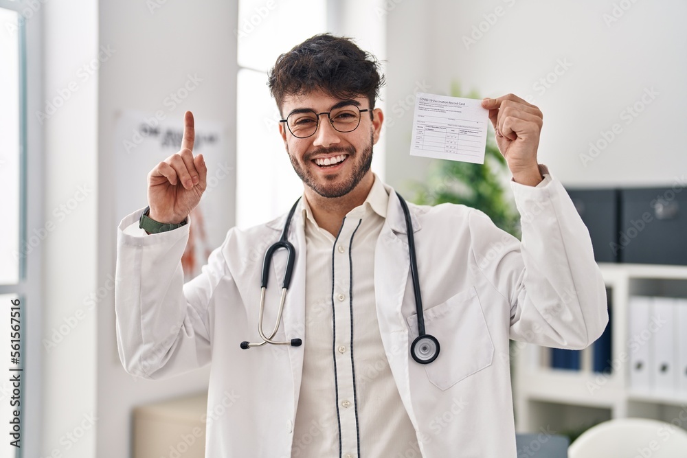 Hispanic doctor man with beard holding covid record card surprised with an idea or question pointing finger with happy face, number one