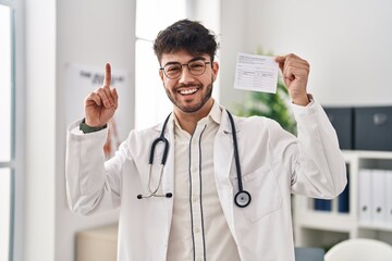 Wall Mural - Hispanic doctor man with beard holding covid record card surprised with an idea or question pointing finger with happy face, number one