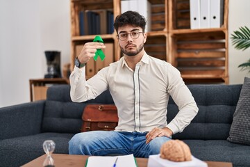 Sticker - Hispanic man with beard holding support green ribbon at therapy clinic thinking attitude and sober expression looking self confident