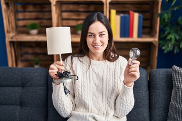 Canvas Print - Young brunette woman holding led lightbulb and lamp smiling with a happy and cool smile on face. showing teeth.