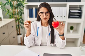 Wall Mural - Young hispanic doctor woman holding heart and cbd oil skeptic and nervous, frowning upset because of problem. negative person.