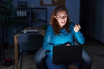 Wall Mural - Brunette woman working at the office at night angry and mad raising fist frustrated and furious while shouting with anger. rage and aggressive concept.