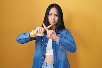 Canvas Print - Young asian woman standing over yellow background rejection expression crossing fingers doing negative sign