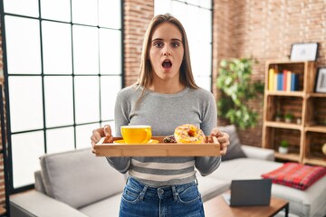 Poster - Young woman holding tray with breakfast food in shock face, looking skeptical and sarcastic, surprised with open mouth