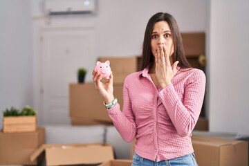 Wall Mural - Young hispanic woman moving to a new home holding piggy bank covering mouth with hand, shocked and afraid for mistake. surprised expression