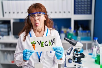 Sticker - Middle age hispanic woman working at vegan laboratory sticking tongue out happy with funny expression.