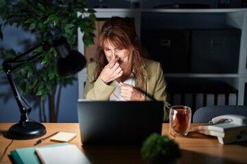 Sticker - Middle age hispanic woman working using computer laptop at night smelling something stinky and disgusting, intolerable smell, holding breath with fingers on nose. bad smell