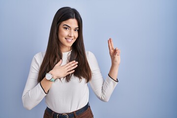 Sticker - Young brunette woman standing over blue background smiling swearing with hand on chest and fingers up, making a loyalty promise oath