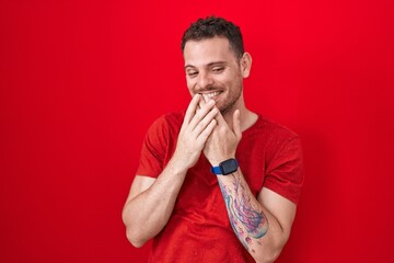 Sticker - Young hispanic man standing over red background laughing and embarrassed giggle covering mouth with hands, gossip and scandal concept