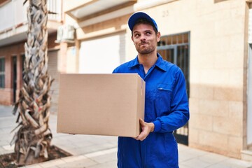 Poster - Young hispanic man delivering box smiling looking to the side and staring away thinking.