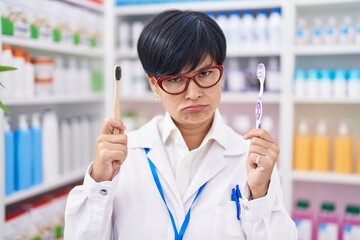 Sticker - Young asian woman with short hair doing toothbrush comparative at pharmacy skeptic and nervous, frowning upset because of problem. negative person.