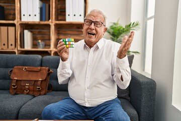 Canvas Print - Senior therapist with grey hair playing colorful puzzle cube intelligence game celebrating achievement with happy smile and winner expression with raised hand