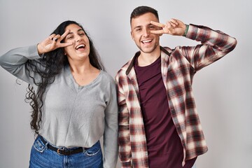 Poster - Young hispanic couple standing over white background doing peace symbol with fingers over face, smiling cheerful showing victory