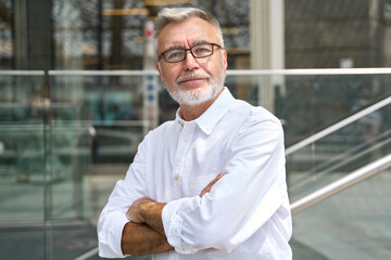 Confident happy mature older business man leader, smiling middle aged senior old professional businessman wearing white shirt glasses crossed arms looking at camera standing outside, portrait.