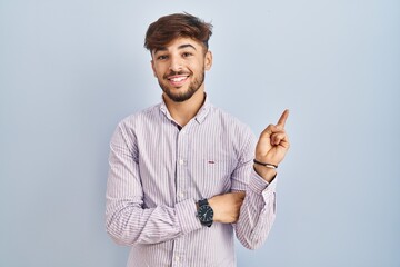 Poster - Arab man with beard standing over blue background with a big smile on face, pointing with hand finger to the side looking at the camera.