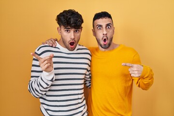 Wall Mural - Young hispanic gay couple standing over yellow background surprised pointing with finger to the side, open mouth amazed expression.