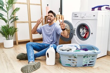 Wall Mural - Young hispanic man putting dirty laundry into washing machine smiling with tongue out showing fingers of both hands doing victory sign. number two.