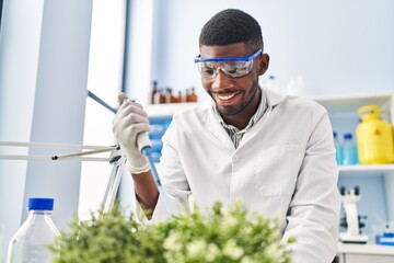 Canvas Print - Young african american man wearing scientist uniform using pipette on plant at laboratory