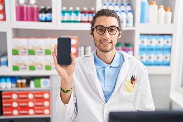 Poster - Young hispanic man working at pharmacy drugstore showing smartphone screen looking positive and happy standing and smiling with a confident smile showing teeth