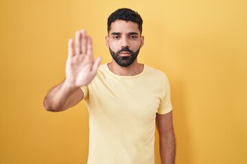 Poster - Hispanic man with beard standing over yellow background doing stop sing with palm of the hand. warning expression with negative and serious gesture on the face.