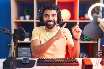 Wall Mural - Hispanic man with beard playing video games with headphones smiling and looking at the camera pointing with two hands and fingers to the side.