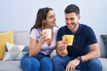 Wall Mural - Young hispanic couple drinking coffee sitting on sofa at home