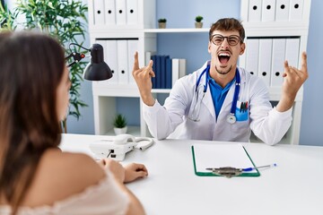 Sticker - Young doctor with client at medical clinic shouting with crazy expression doing rock symbol with hands up. music star. heavy concept.