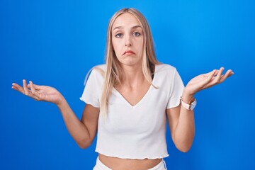 Poster - Young caucasian woman standing over blue background clueless and confused expression with arms and hands raised. doubt concept.