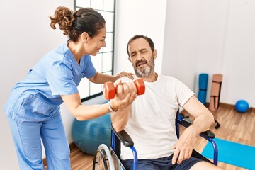 Wall Mural - Middle age man and woman having rehab session using dumbbell sitting on wheelchair at physiotherapy clinic